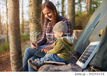 Mother with son on camping vacation look at mobile phone while enjoying the fresh air near tent and solar panel Mother with son on camping vacation look at mobile phone while enjoying the fresh air near tent and solar panel 64354159