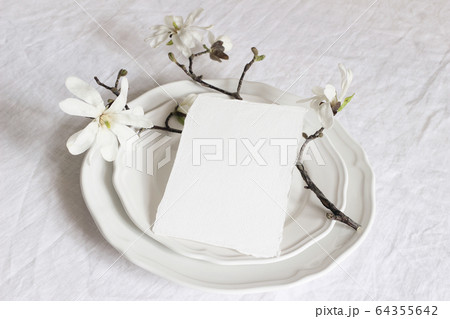 Festive table spring setting with porcelain plates, magnolia stellata tree branches with white blossoms and blank paper card mockup. Feminine composition, wedding or restaurant menu concept. 64355642