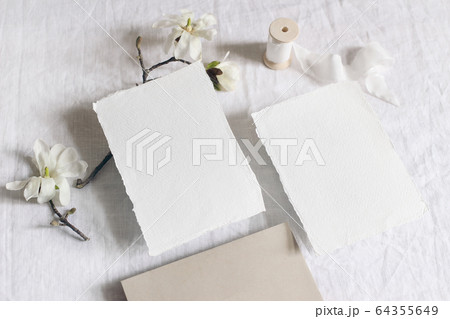 Wedding stationery set. Mock-up scene with blank paper greeting cards, envelope on linen tablecloth background. White magnolia stellata tree branches and ribbon. Feminine still life, flat lay, top. 64355649