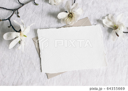 Wedding stationery mock-up scene. Blank horizontal greeting card, envelope on linen tablecloth background with white magnolia stellata tree branches and ribbon. Feminine still life. Flat lay, top view 64355669