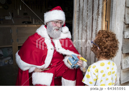 Man wearing Santa Claus costume giving wooden toy car to boy with red curly hair. 64365091