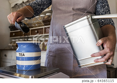 Close up of woman standing in a kitchen, placing loose tea into striped blue ceramic jar. Close up of woman standing in a kitchen, placing loose tea into striped blue ceramic jar. 64365115