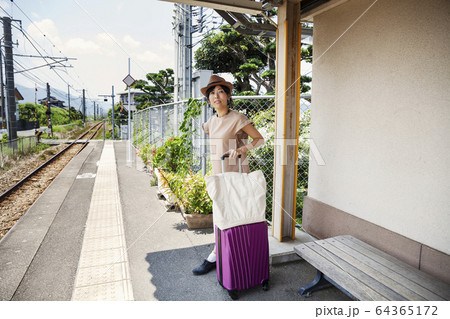 Japanese woman wearing hat standing on railway station platform with shopping bag and pink suitcase. 64365172