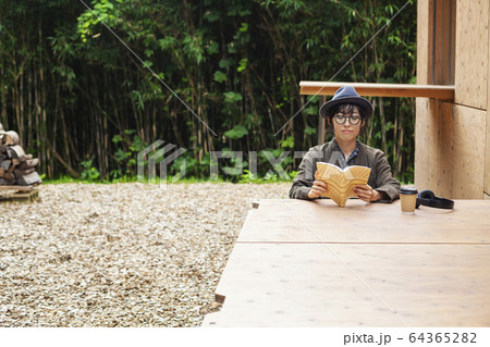Japanese woman wearing glasses and hat sitting at a table outside Eco Cafe, reading book. 64365282
