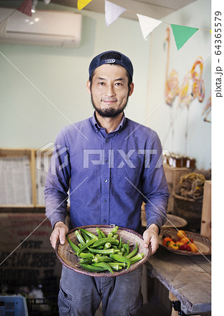 Smiling Japanese man wearing cap standing in farm shop, holding bowl with fresh okra. 64365579