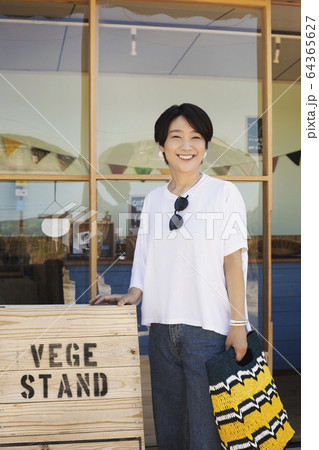 Japanese woman standing outside a farm shop, holding shopping bag, smiling at camera. 64365627