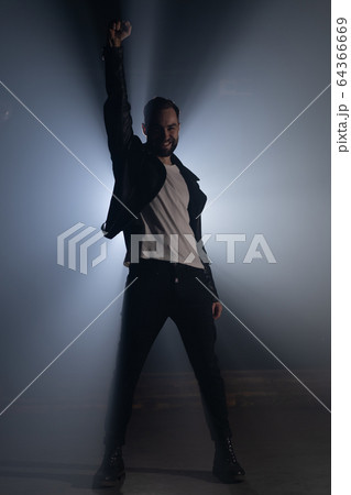 Young man in leather jacket on smoky stage. View of guy raises hand up as gesture and symbol of 64366669