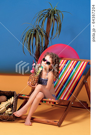 Smiling girl in swimsuit and sunglasses sitting in rainbow deck chair holding pineapple cocktail with colorful straws and showing thumb up and sunbathing Smiling girl in swimsuit and sunglasses sitting in rainbow deck chair holding pineapple cocktail with colorful straws and showing thumb up and sunbathing 64367204