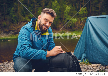 A young, bearded man with a backpack, camping, outdoors, against the backdrop of a tourist tent. A young, bearded man with a backpack, camping, outdoors, against the backdrop of a tourist tent. 64371551