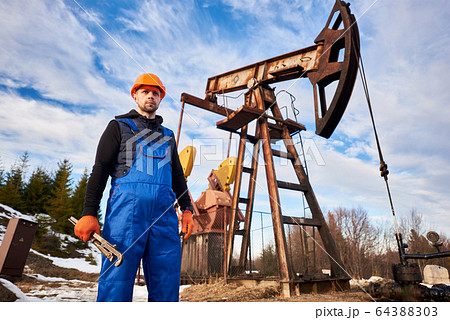 Young oil man standing in front of pump jack. Young oil man standing in front of pump jack. 64388303