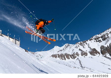 A male athlete skier in an orange trigger makes a jump jump with a grab with flying snow powder against the background of Mount Elbrus in the North Caucasus. Winter Extreme Sports Concept 64405909