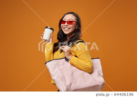 Asian woman looking at camera in pink glasses with bag on empty yellow background Asian woman looking at camera in pink glasses with bag on empty yellow background 64406196