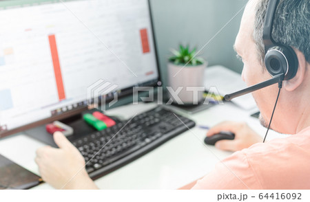 Closeup of man back view in microphone headset sitting at the computer and white desktop. Staying at home, working remotely, distance customer service, call center, home office, help desk concept Closeup of man back view in microphone headset sitting at the computer and white desktop. Staying at home, working remotely, distance customer service, call center, home office, help desk concept 64416092