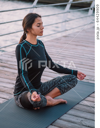 Young brunette woman doing yoga exercises on wooden sea embankment in the morning. Girl in black 64429909