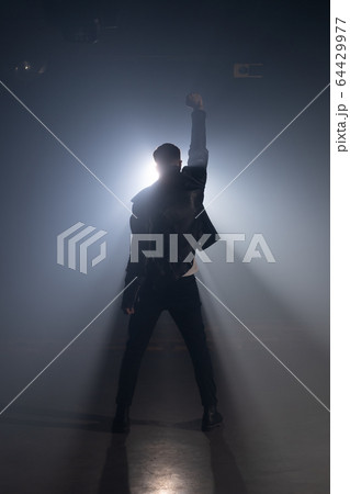 Young man in leather jacket on smoky stage. View of guy raises hand up as gesture and symbol of Young man in leather jacket on smoky stage. View of guy raises hand up as gesture and symbol of 64429977