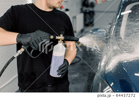 Car wash with flowing water and foam. Side view of young car wash service male employee holding high pressure spray with cleaning foam. Carwash. Washing machine at the station 64437272
