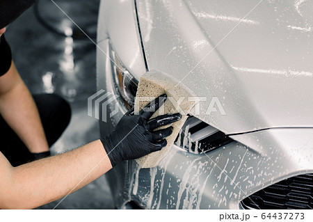 Manual washing of a car with soap and sponge. Cropped image of hand of young man worker in protective clothes, holding a sponge for washing car headlight 64437273