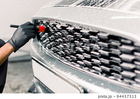 Car detailing wash. Detailing cleaning with soap at car wash. Cropped image of hand of male worker in rubber gloves washing car radiator grille of luxury white car with special brush 64437313