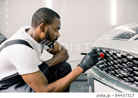 Close up photo of young African male worker in black protective gloves cleaning radiator grille in foam with special brush. Carwash and detailing. Washing machine at the station. Car washing concept. 64437317
