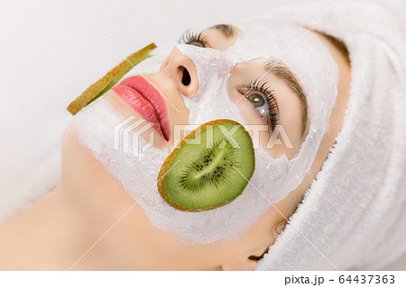 Facial treatment, skin care, home spa. Close up side view photo of young woman in towel on her head, with natural homemade fruit facial mask on skin and slices of fresh kiwi on cheeks. 64437363