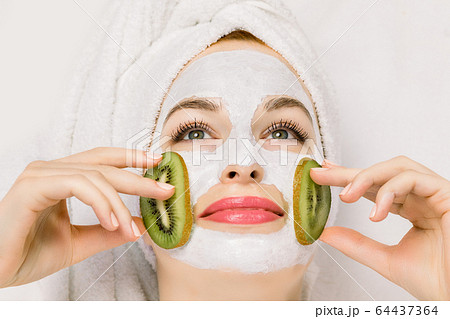 Close up of pretty smiling woman with white moisturizing mask and pieces of fresh kiwi fruit on cheeks. Beauty facial mask, skin care and treatment concept Close up of pretty smiling woman with white moisturizing mask and pieces of fresh kiwi fruit on cheeks. Beauty facial mask, skin care and treatment concept 64437364