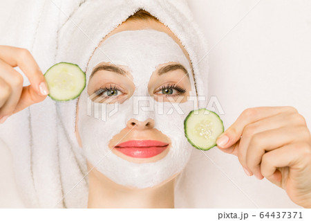 Home spa, skin care concept. Close up top portrait of pretty young woman with towel on her head, white clay mask on her face, holding pieces of fresh cucumber, lying on the isolated white background. 64437371