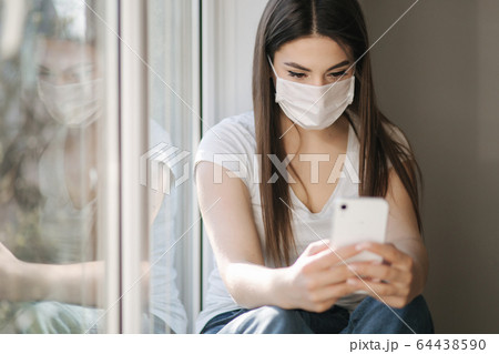 Attractive woman in medical mask sits on windowsill at home. Female in white t'shirt and white medical mask use phone 64438590