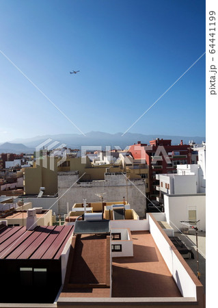 Charter plane leaving Tenerife airport during sunrise seen from roofs of traditional houses with Mount Teide volcano in background, Canary Islands, Spain, sunny blue sky with copy space 64441199