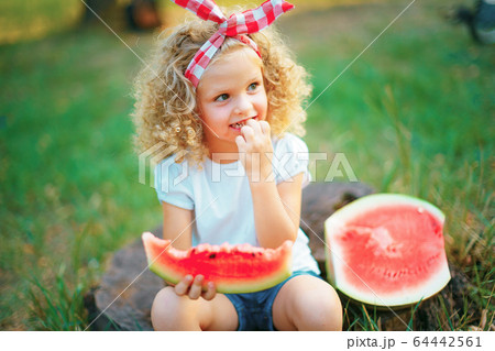 Happy curly child girl sitting on stump and eating watermelon outdoors in spring park against natural sunny blurred background Happy curly child girl sitting on stump and eating watermelon outdoors in spring park against natural sunny blurred background 64442561