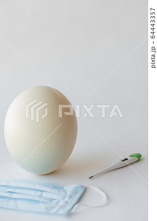 Large white easter ostrich egg, medical mask and thermometer. Egg, respirator and thermometer. Easter and Coronavirus, Covid-19. Ostrich egg, medical mask and thermometer on a white background. 64443357