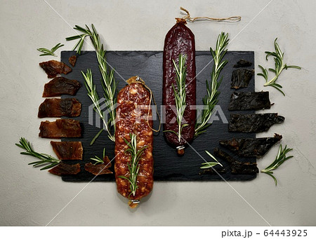Raw smoked and cured wild meat products with rosemary on a black backing on a gray background in a cool shade. 64443925