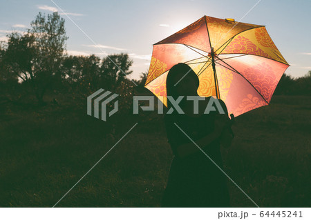 A girl with a colored umbrella poses in a field. The umbrella is illuminated from behind by the sunset light 64445241