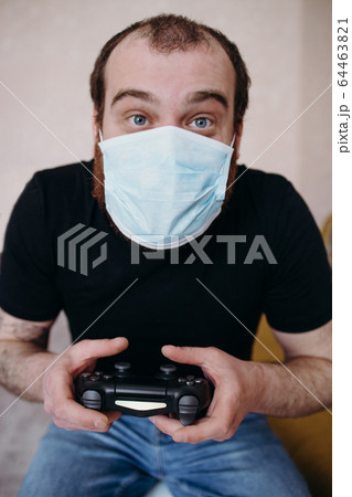 Adult balding white man playing video games with medical mask on his face while Covid-19 quarantine Adult balding white man playing video games with medical mask on his face while Covid-19 quarantine 64463821