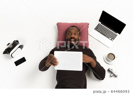 Emotional african-american man using tablet surrounded by gadgets isolated on white studio background, technologies connecting people. Shocked, scared 64466093