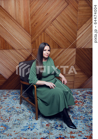 Pretty young lady in a evening dress sitting in a chair on wooden background. Plus size caucasian female model posing in beautiful evening gown 64474004