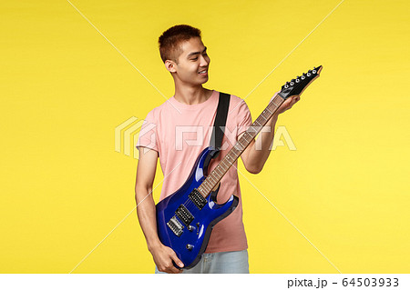 Lifestyle, leisure and youth concept. Cheerful handsome asian male student playing in band, tune electric guitar and smiling pleased, ready to jam on stage, perform new song, yellow background 64503933