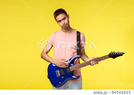 Lifestyle, leisure and youth concept. Gloomy asian guy playing in band, look sad and uneasy, have bad day, holding electric guitar, trying learn new instrument, stand yellow background Lifestyle, leisure and youth concept. Gloomy asian guy playing in band, look sad and uneasy, have bad day, holding electric guitar, trying learn new instrument, stand yellow background 64503935