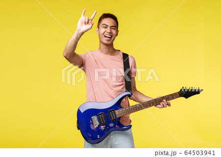 Lifestyle, leisure and youth concept. Happy carefree, good-looking asian guy playing in band, hold electric guitar, show rock-n-roll heavy metal sign and smiling broadly, perform on stage 64503941