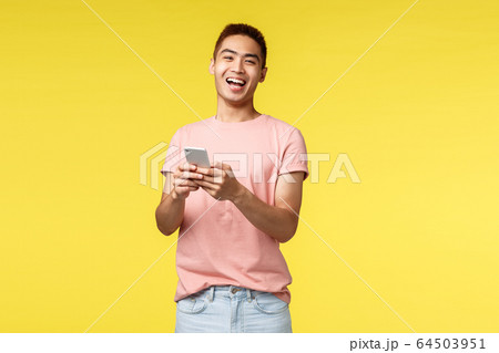 Technology, communication and lifestyle concept. Enthusiastic asian male in pink t-shirt laughing from cool joke or video, using mobile phone, smiling pleased camera, stand yellow background Technology, communication and lifestyle concept. Enthusiastic asian male in pink t-shirt laughing from cool joke or video, using mobile phone, smiling pleased camera, stand yellow background 64503951