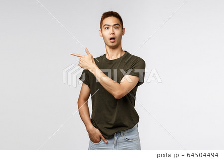 Portrait of surprised, curious asian male staring camera startled, asking question and pointing finger left at interesting strange item in store, showing way, standing grey background Portrait of surprised, curious asian male staring camera startled, asking question and pointing finger left at interesting strange item in store, showing way, standing grey background 64504494