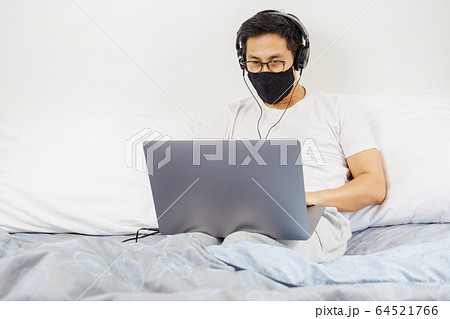 Man with protective mask and headphone working on laptop in bed at home covid-19 outbreak. 64521766