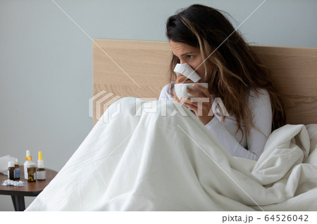 Sick young woman blowing nose, holding handkerchief, sitting in bed Sick young woman blowing nose, holding handkerchief, sitting in bed 64526042
