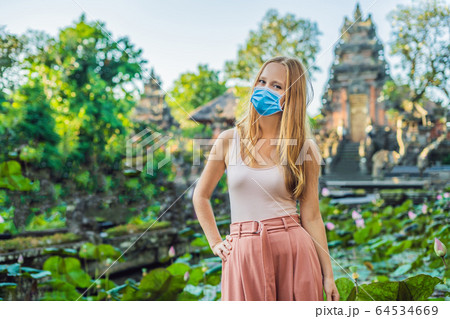Young woman in medical mask traveler in the background of Pura Taman Kemuda Saraswati Temple in Ubud 64534669