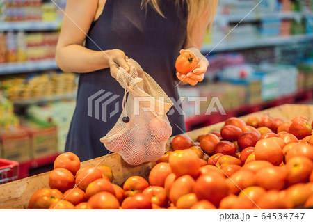 A woman chooses tomatoes in a supermarket without using a plastic bag. Reusable bag for buying 64534747