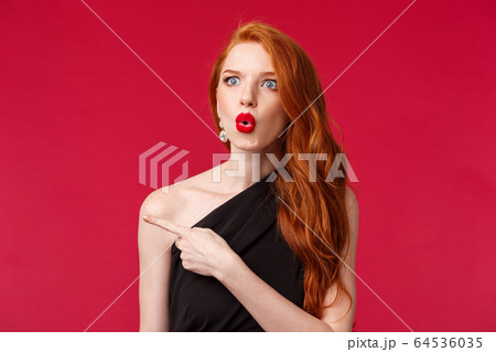 Close-up portrait of impressed excited and speechless attractive redhead woman, wearing black dress, red lipstick and evening makeup. look startled say wow pointing left questioned 64536035