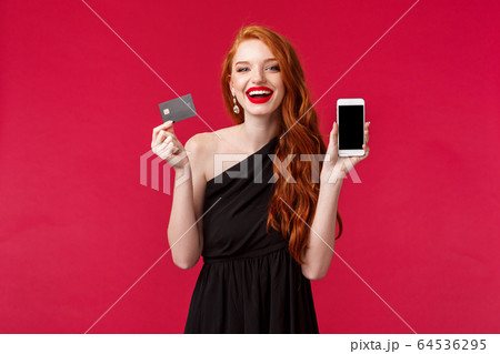 Portrait of gorgeous charismatic redhead woman in black dress, showing mobile phone display and credit card, paying for stunning black dress online in internet store, look satisfied smiling 64536295