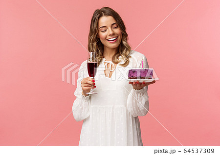 Holidays, spring and party concept. Portrait of tender, elegant young blond woman holding glass of wine and cake with lit candle, smiling pleased, celebrating birthday, pink background 64537309