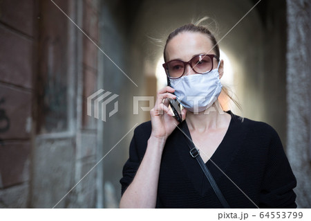 COVID-19 pandemic coronavirus. Casual caucasian woman at medieval city street using mobile phone, wearing protective face mask against spreading of coronavirus and disease transmission COVID-19 pandemic coronavirus. Casual caucasian woman at medieval city street using mobile phone, wearing protective face mask against spreading of coronavirus and disease transmission 64553799