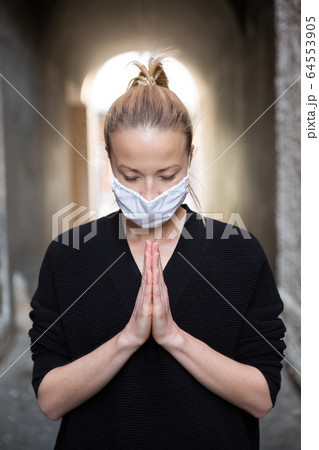 Coronavirus outbreak. Young caucasian woman wearing medical protection face mask praying over coronavirus global pandemic, for salvation of humanity, health, anxiety and depression reduction 64553905
