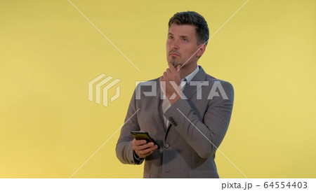 Tall man in suit holding one hand on chin, looking on smartphone and thinking about something. Tall man in suit holding one hand on chin, looking on smartphone and thinking about something. 64554403